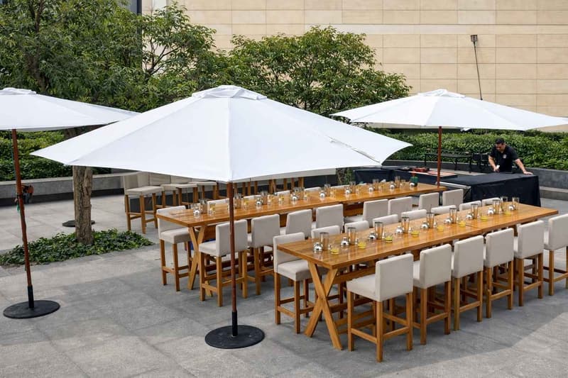Montaje de comedor tipo lounge en exterior con mesas altas de madera, bancos tapizados y sombrillas de gran formato para eventos corporativos o sociales en espacio abierto