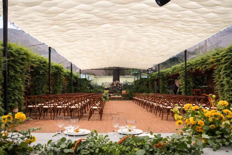 Montaje de ceremonia en exterior con pasillo central, sillas tipo crossback en disposición simétrica y cubierta textil ondulada para control de luz, en ambiente natural con decoración floral