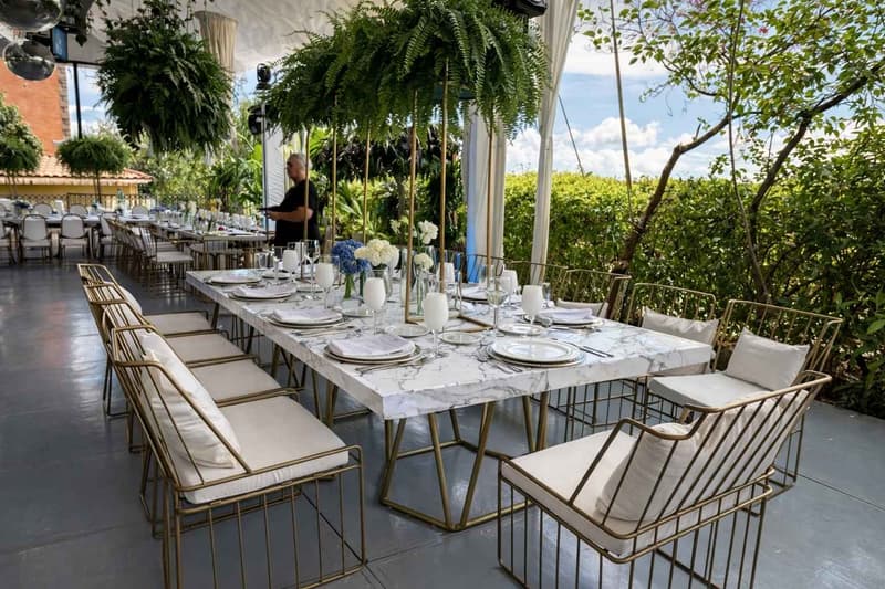 Montaje de comedor elegante en exterior con mesa tipo mármol, sillas metálicas con cojinería clara y ambientación natural con follaje colgante para eventos sociales o corporativos de alto nivel